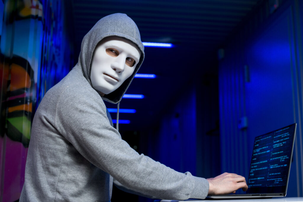 Man with red light therapy mask wearing hoodie while typing on a laptop.